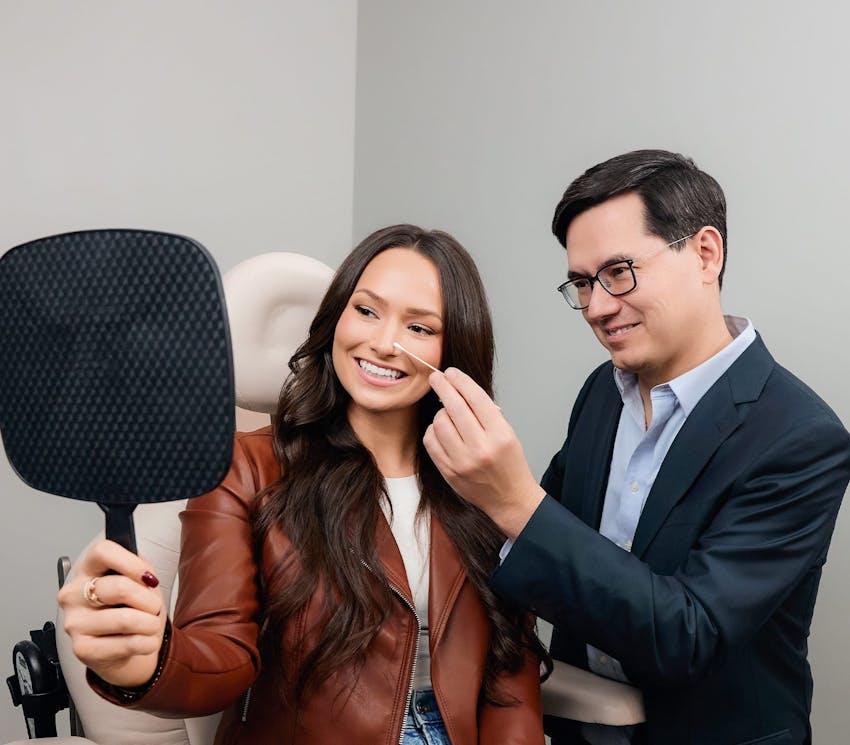 woman smiling in handheld mirror with doctor pointing to her nose