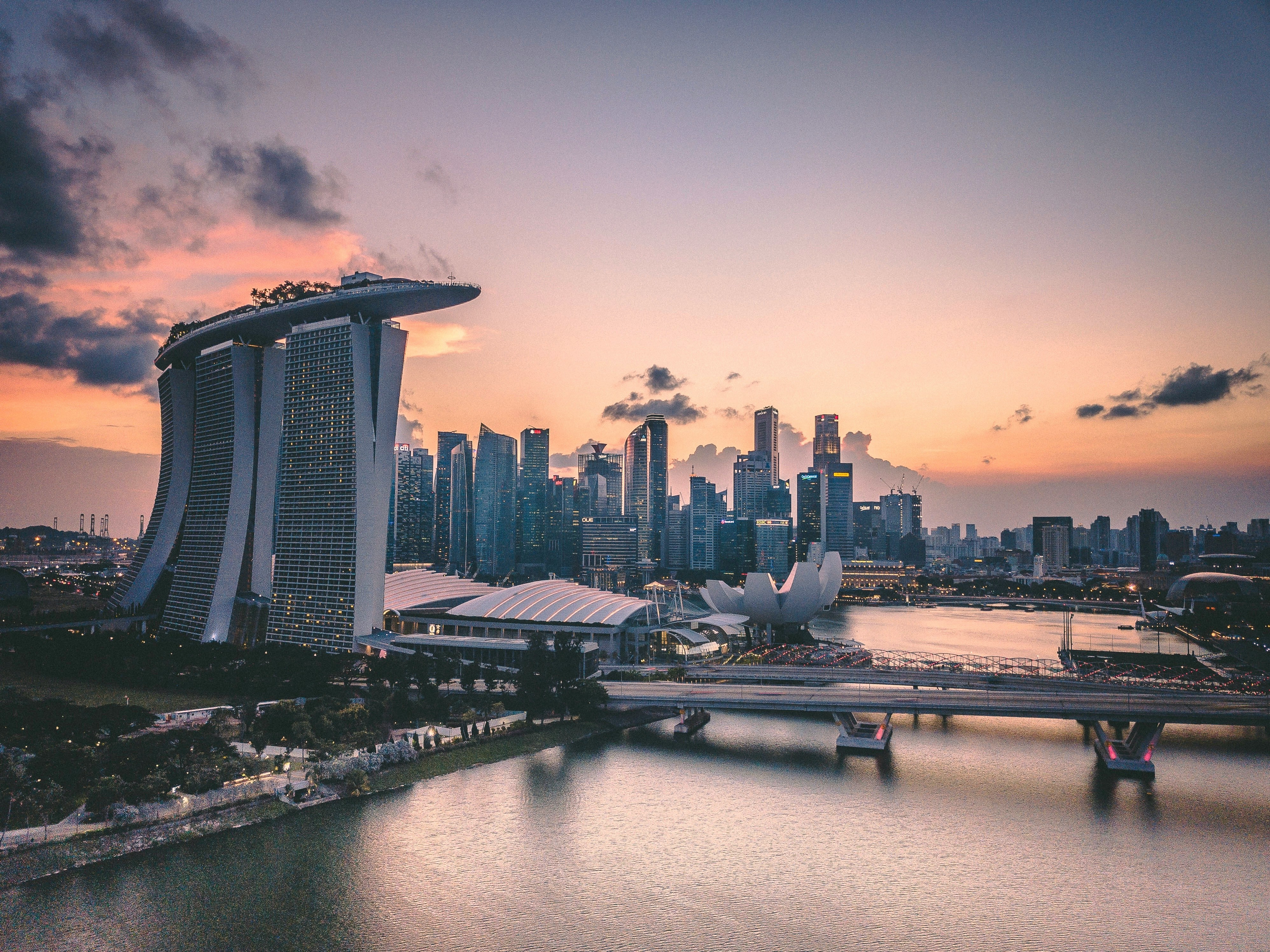 Aerial view of Singapore skyline at sunset, featuring Marina Bay Sands hotel, the ArtScience Museum, modern skyscrapers, and waterfront with a soft orange and purple sky in the background.
