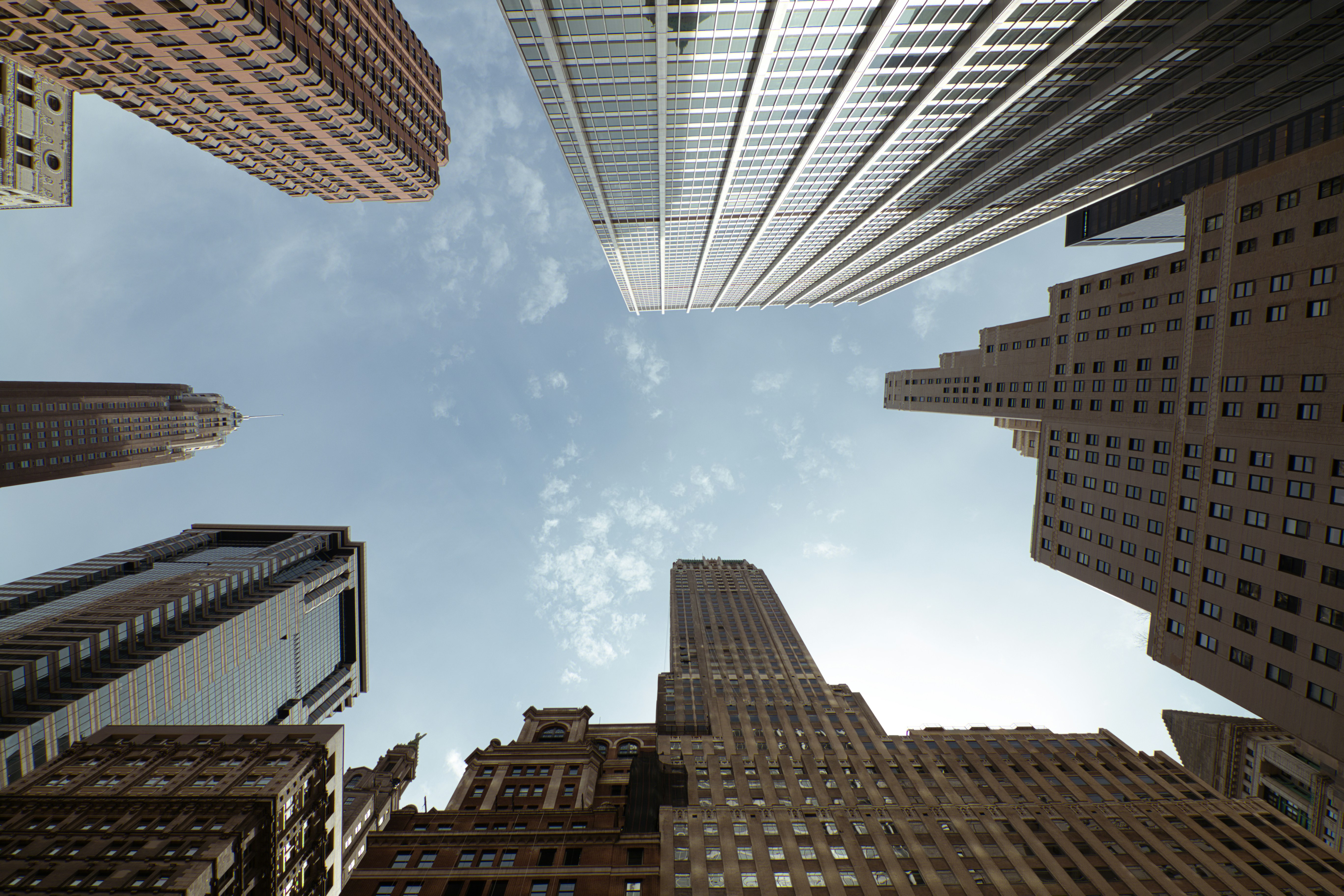 A view looking up at tall skyscrapers from street level in a city, with blue sky and scattered clouds visible between the buildings.