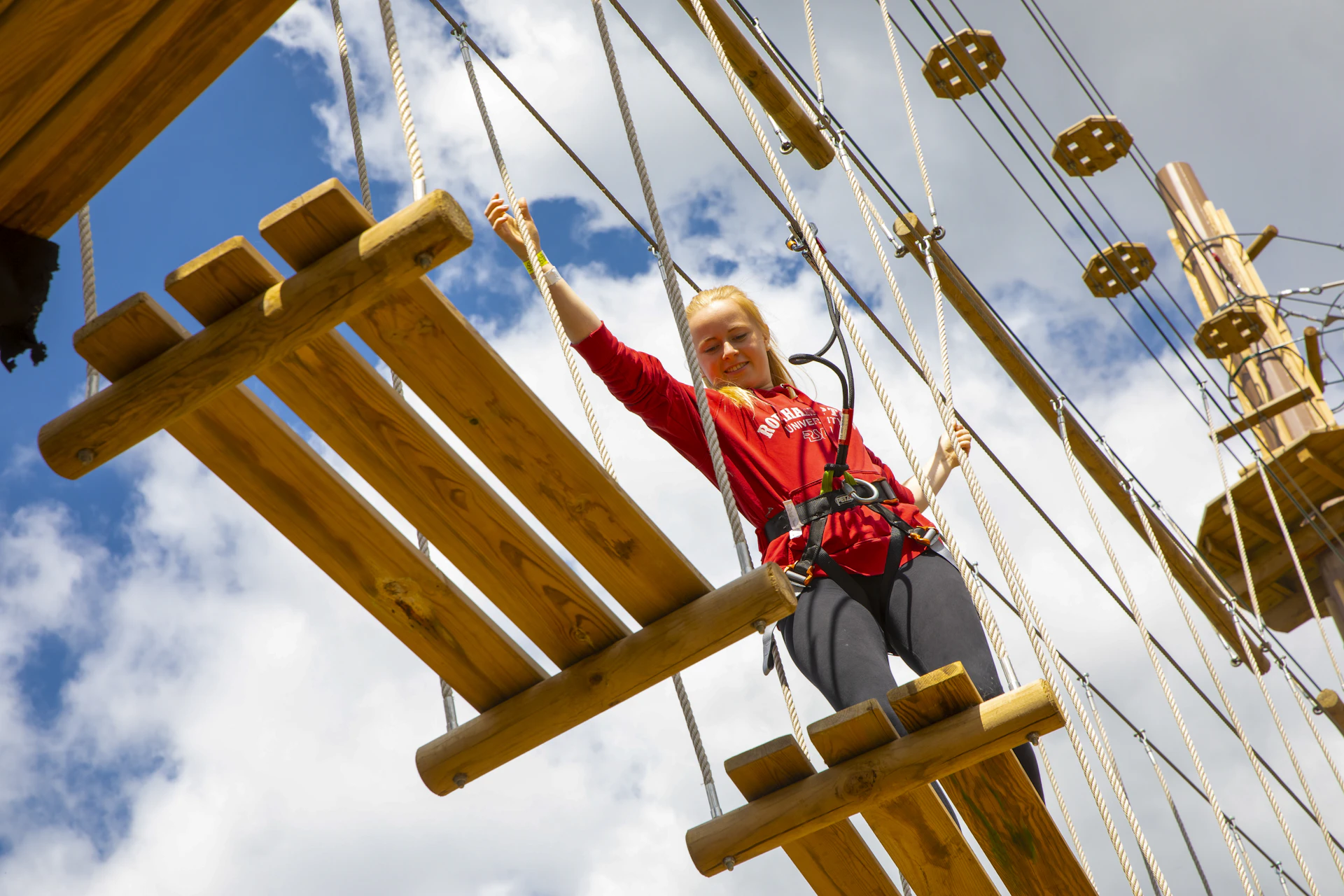 Person high above steps across wooden platforms while wearing a harness.