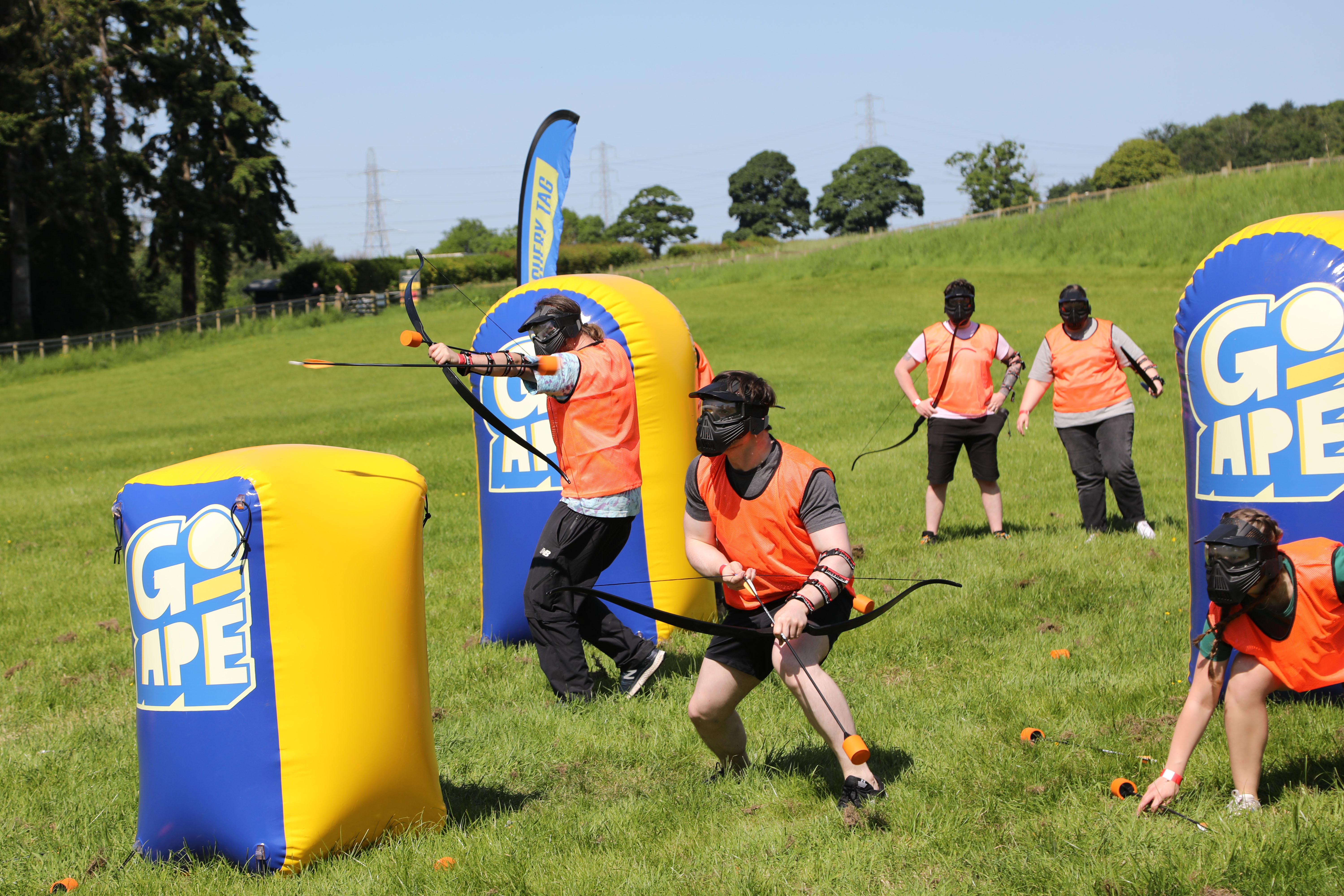 People playing Archery Tag in safety gear using bow and foam-tipped arrows while hiding behind inflatable obstacles.