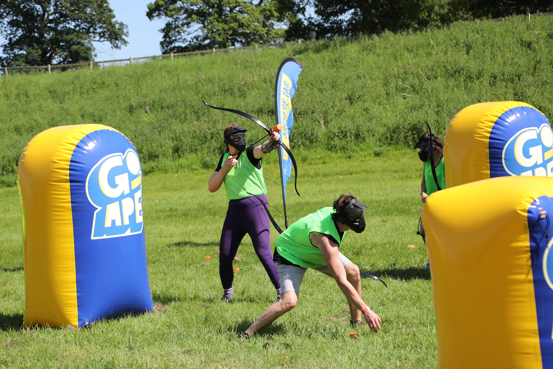 People playing Archery Tag in safety gear using bow and foam-tipped arrows while hiding behind inflatable obstacles.