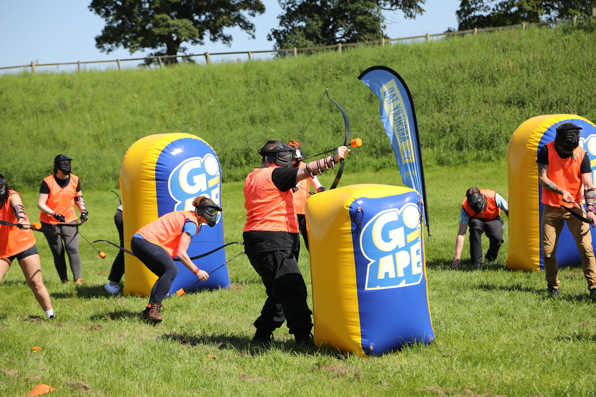 People playing Archery Tag in safety gear using bow and foam-tipped arrows while hiding behind inflatable obstacles.