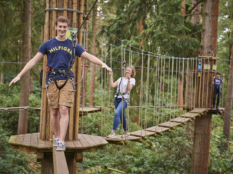 A teenage boy balances on a Go Ape crossing whilst his group complete sections of the course behind him on a half term day out