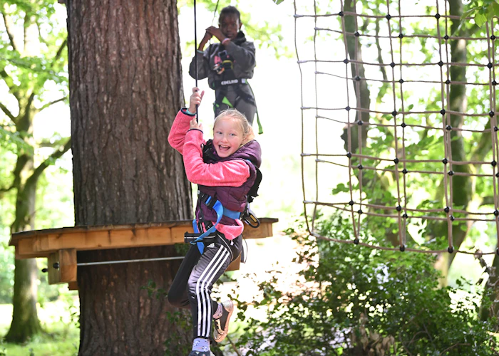 Two young people on a high ropes course, with the person in front on a zip wire while smiling.