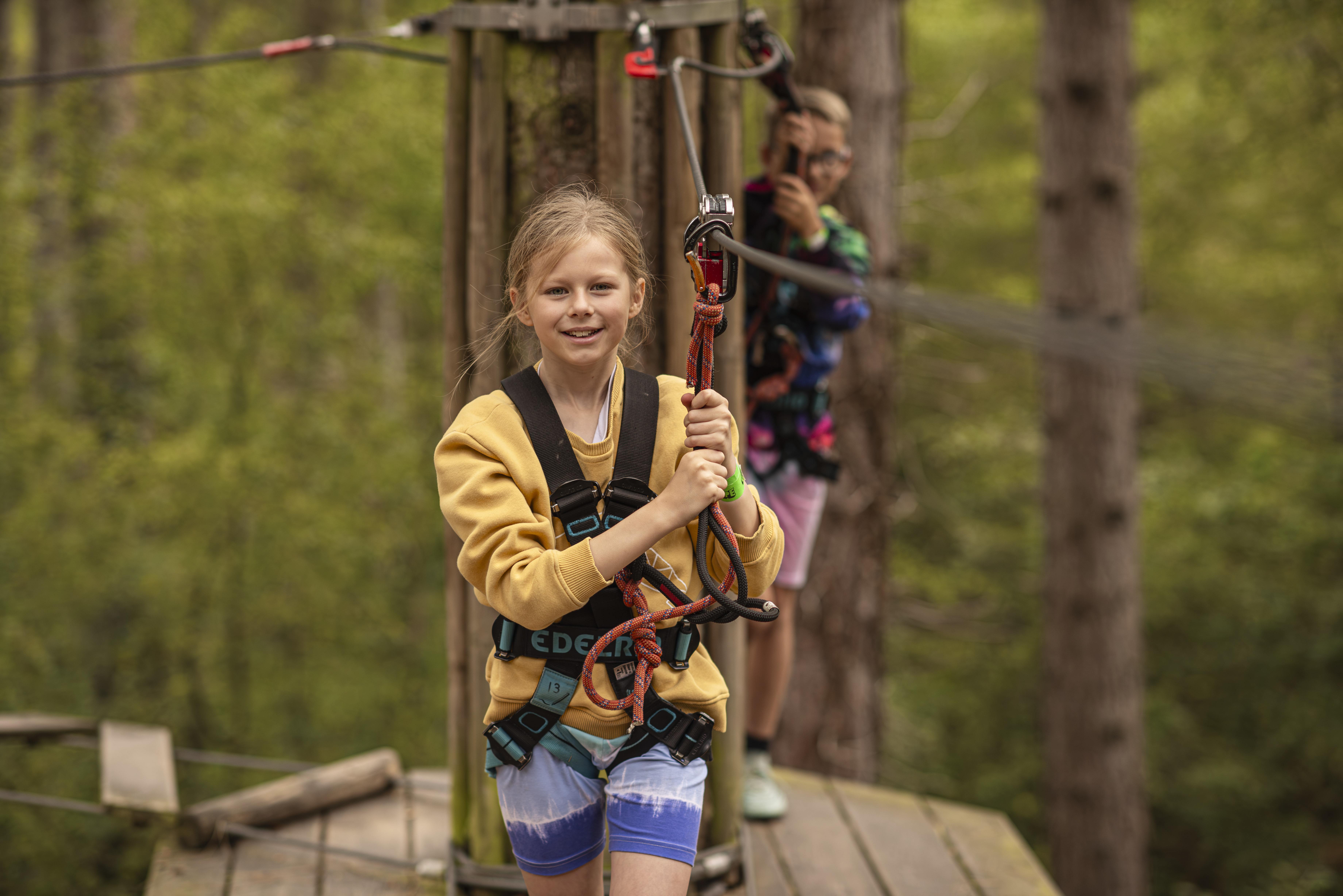 A girl in a yellow jumper crosses a high ropes on her half term day out whilst her friend watches from a platform behind