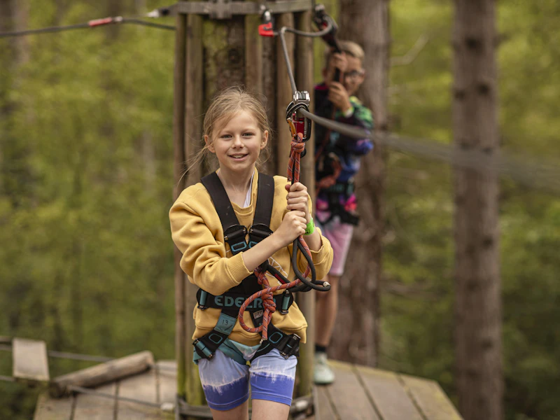 A girl in a yellow jumper crosses a high ropes on her half term day out whilst her friend watches from a platform behind