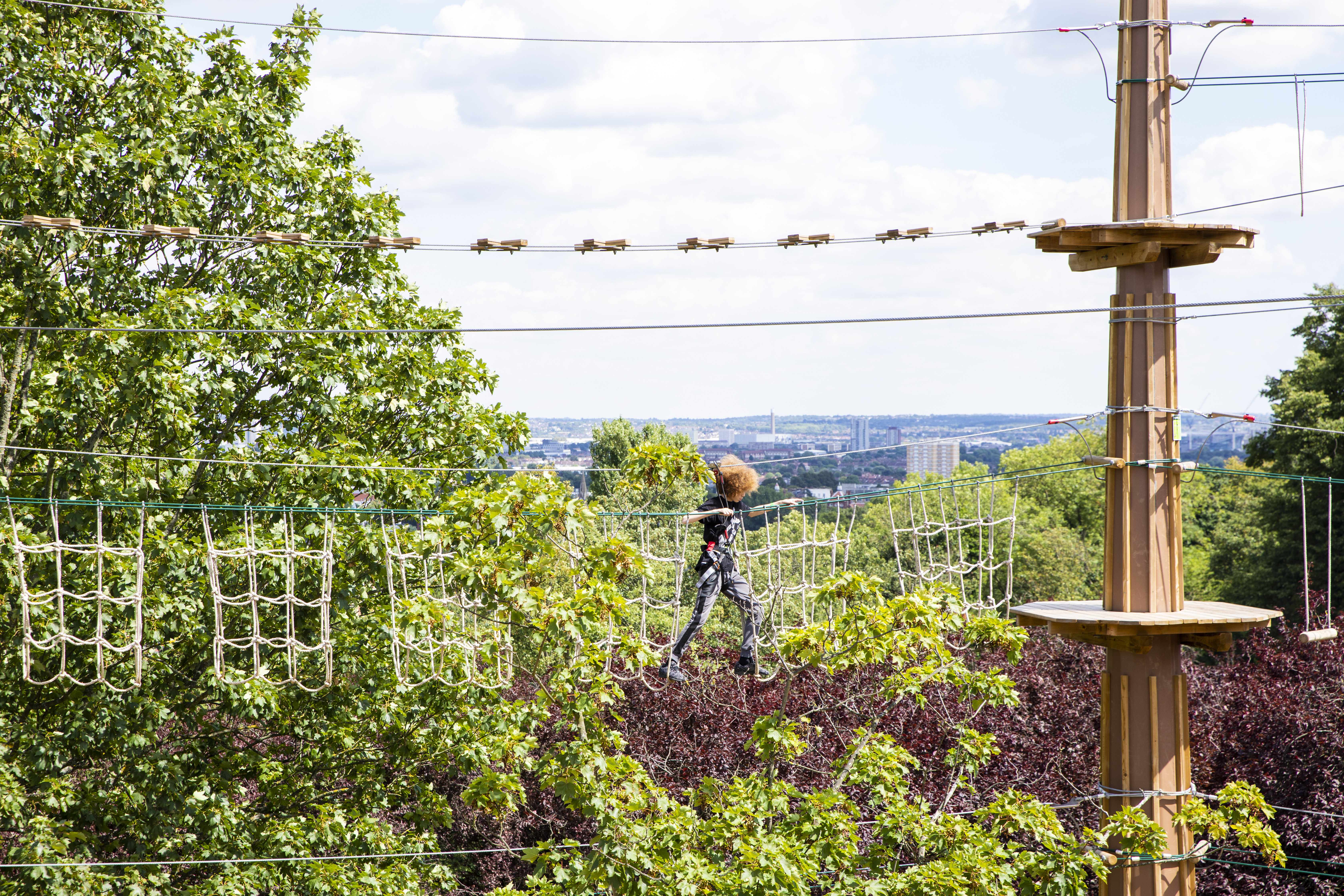 Person steps across a high ropes course with trees and city skyline in the distance.