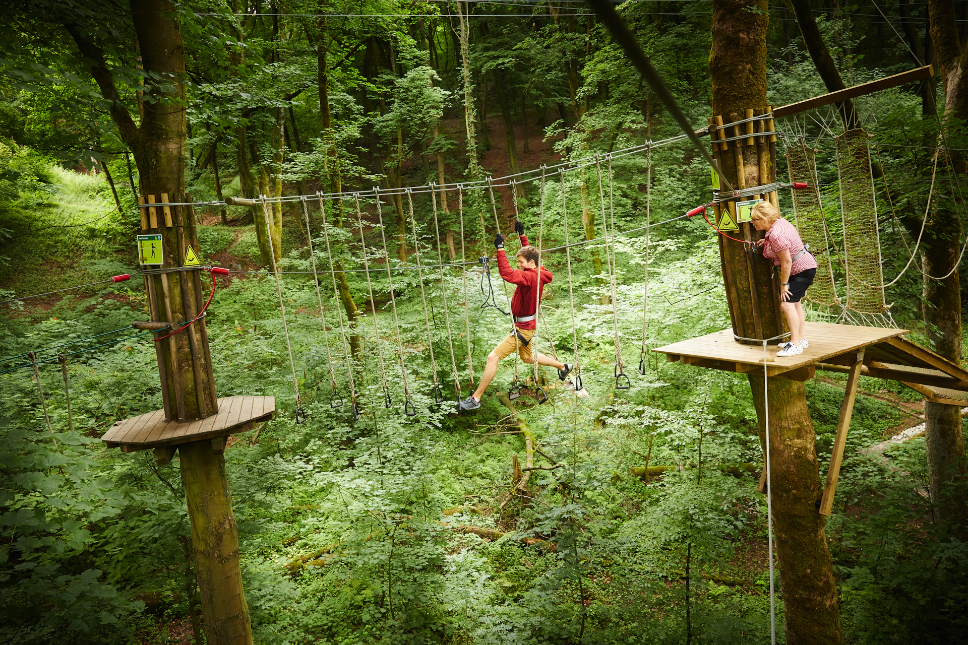 Person on a high ropes crossing while another waits on a platform.