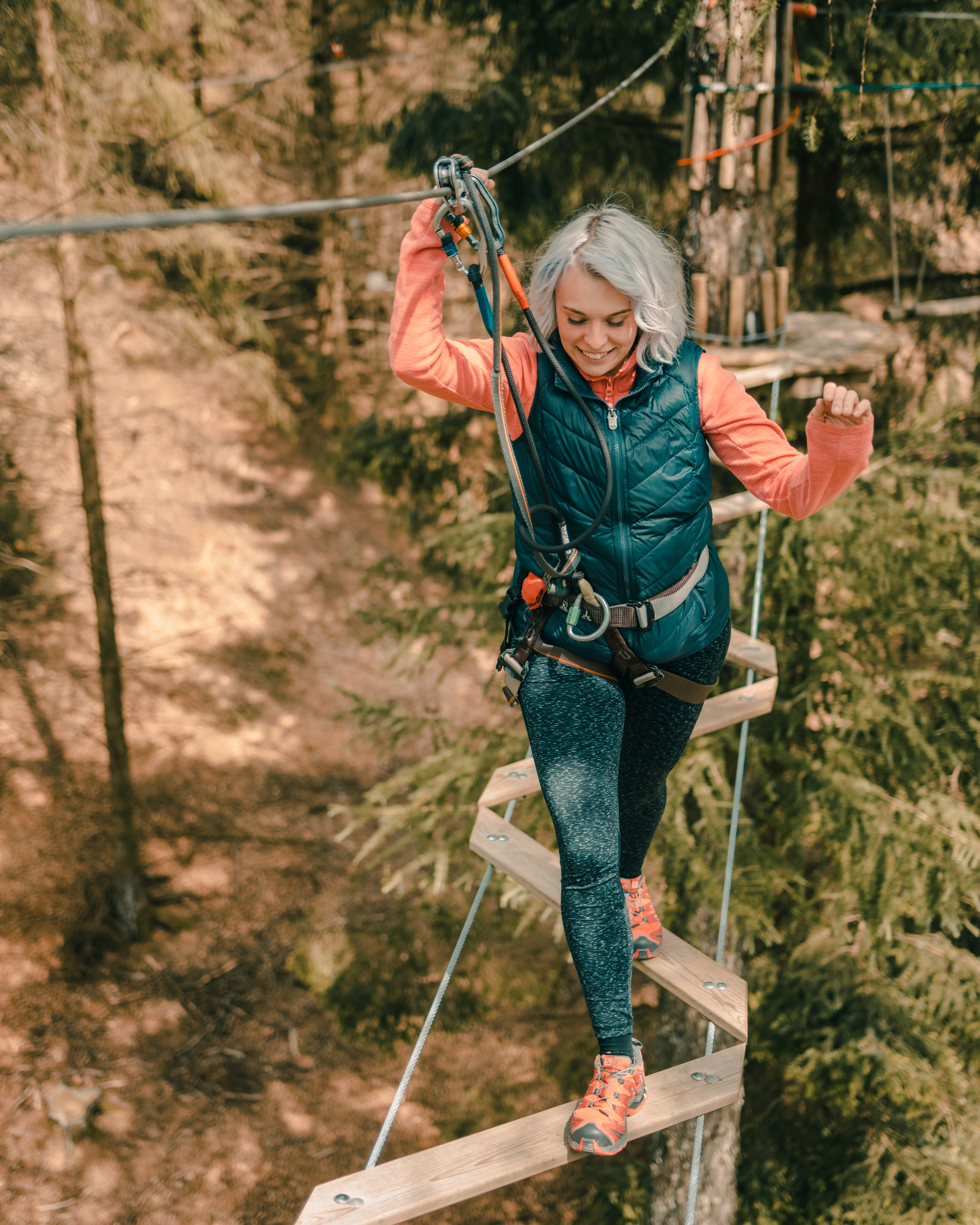 A woman wearing a pink top and gilet smiles as she balances her way over a tree top crossing in the forest