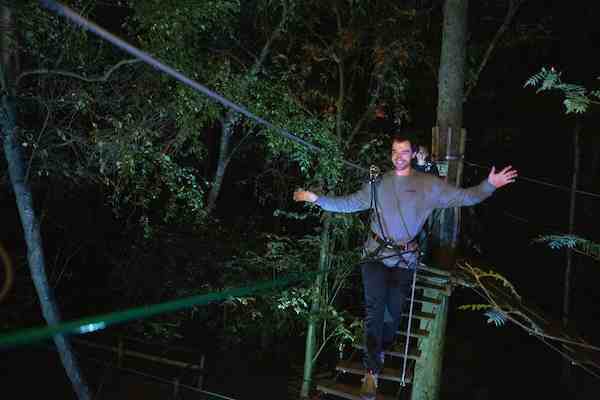 A man grins as he balances his was over a high ropes crossing in the dark