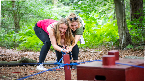 Two women crouch and pull on a length of elastic for a Go Ape team game in the forest
