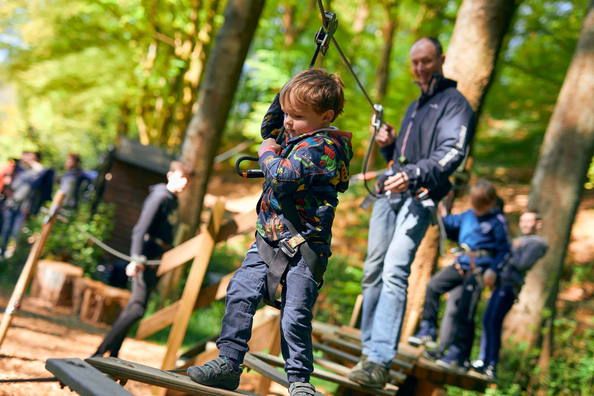 A young boy wearing a coat completes a Go Ape obstacle close tot he ground whilst his did watches on proudly behind him.