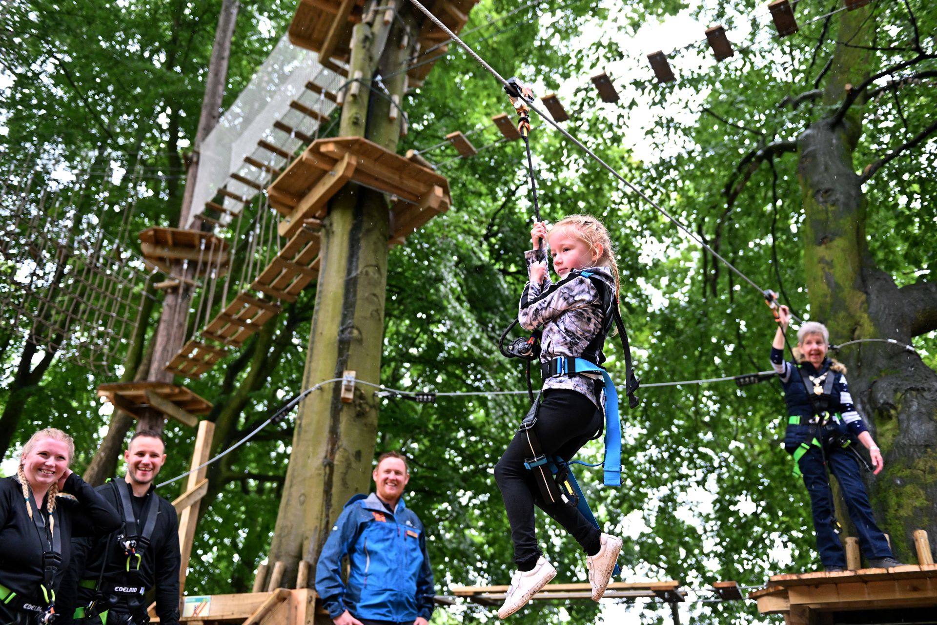 A young girl rides a Go Ape zip wire whilst her supervising adult watches on from a platform behind her, and her parents and a Go Ape instructor cheer her on from the ground.