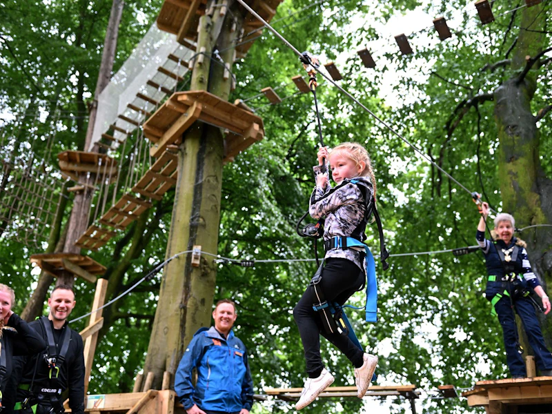 Young girl on zip wire A young girl rides a Go Ape zip wire whilst her supervising adult watches on from a platform behind her, and her parents and a Go Ape instructor cheer her on from the ground.