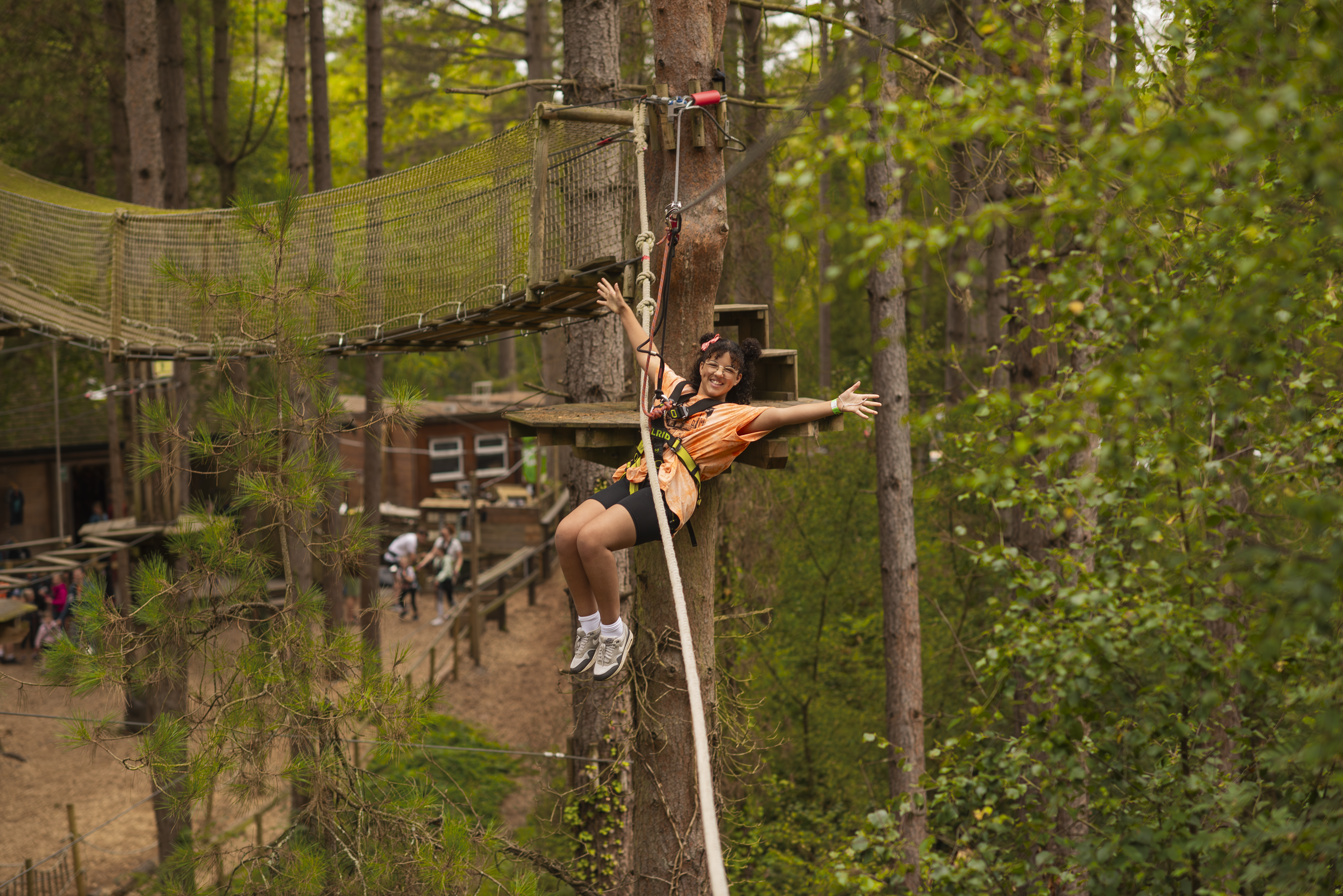 A girl wearing an orange t-shirt throws her arms out as she zips of a high ropes crossing