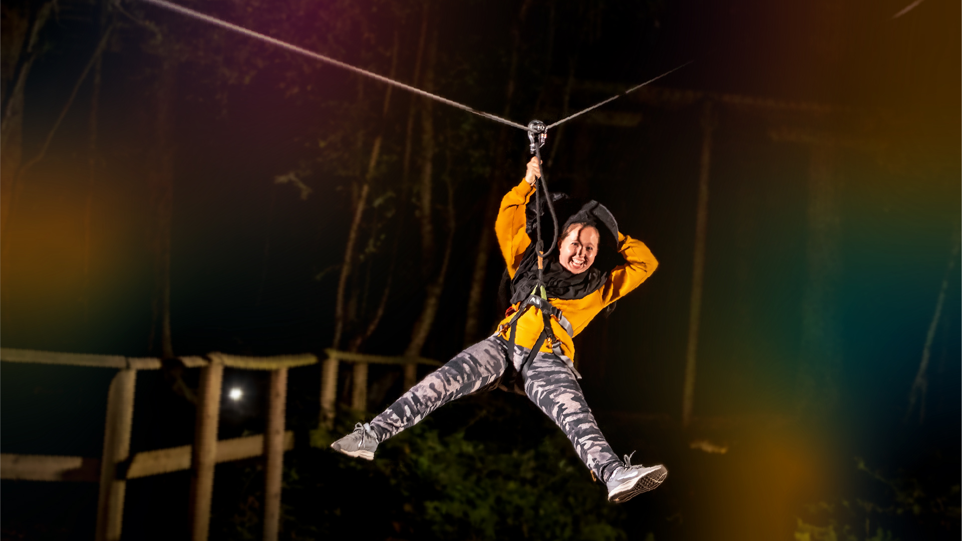 A woman rides a zip wire after dark surrounded bu colourful lights