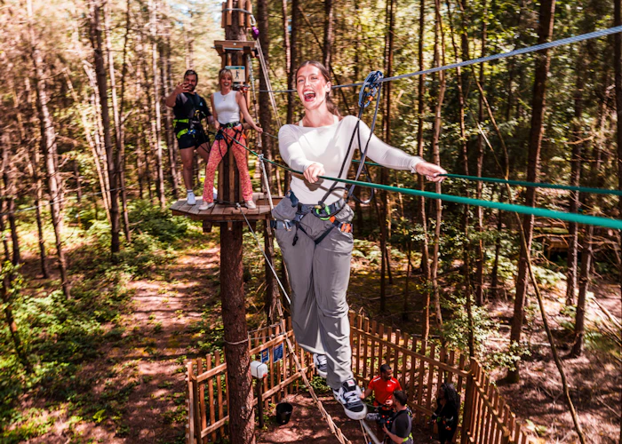 A woman grins as she takes on a Go Ape high ropes crossing whilst her mates watch from the platform behind