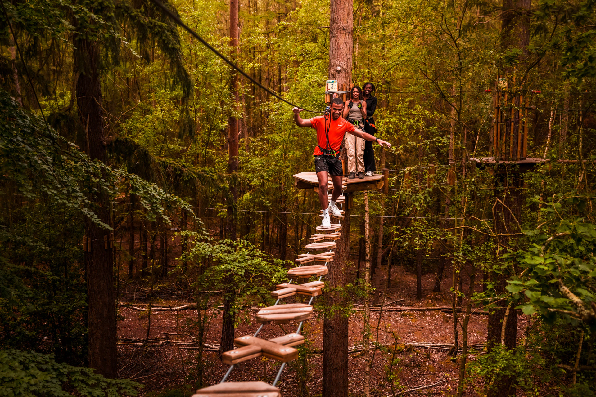 A man balaces on a high ropes crossing whilst his friends watch from a platform behind