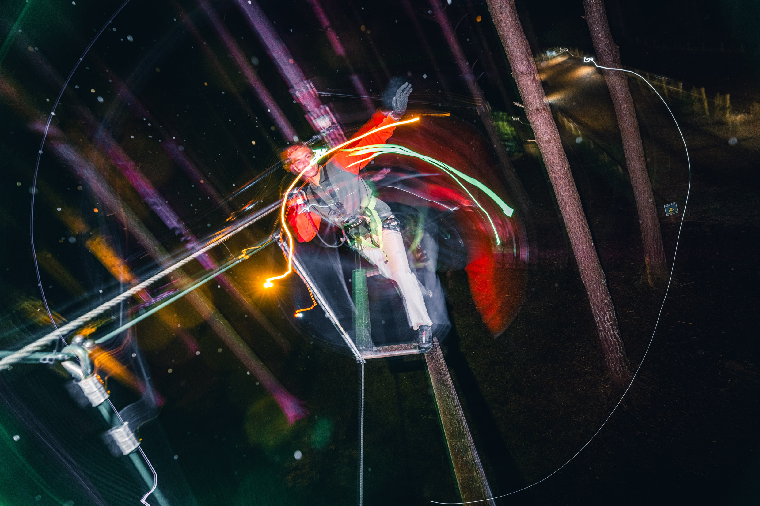 A man balances on a treetop crossing after dark surrrounded by colourful lights