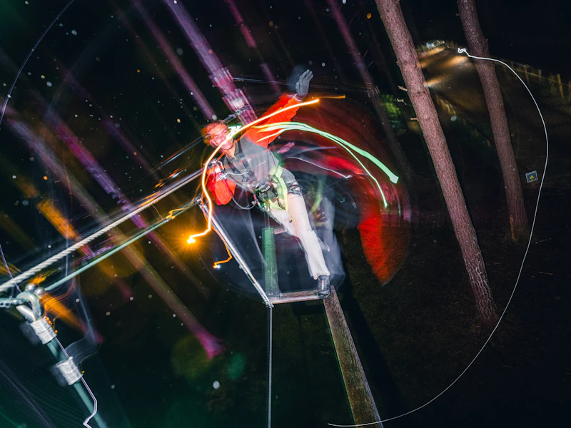 Sundown Adventures Corssing Man A man balances on a treetop crossing after dark surrrounded by colourful lights