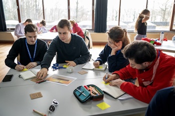 Vier mensen aan tafel die schrijven in een boekje.