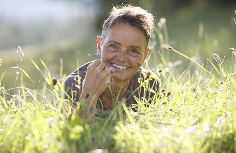 Une femme souriante aux cheveux courts est allongée sur le ventre dans une prairie ensoleillée.