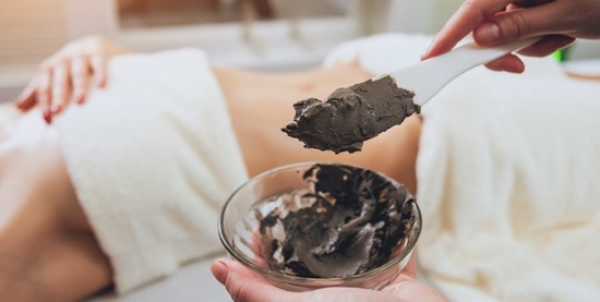 Someone is mixing a brown paste, in the back a woman is lying on a massage table, covered with bath towels