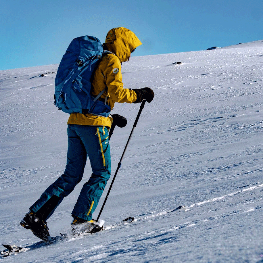 Tourenski Mann im gelben Anorak und mit Wanderstöcken geht einen beschneiten Berg hoch