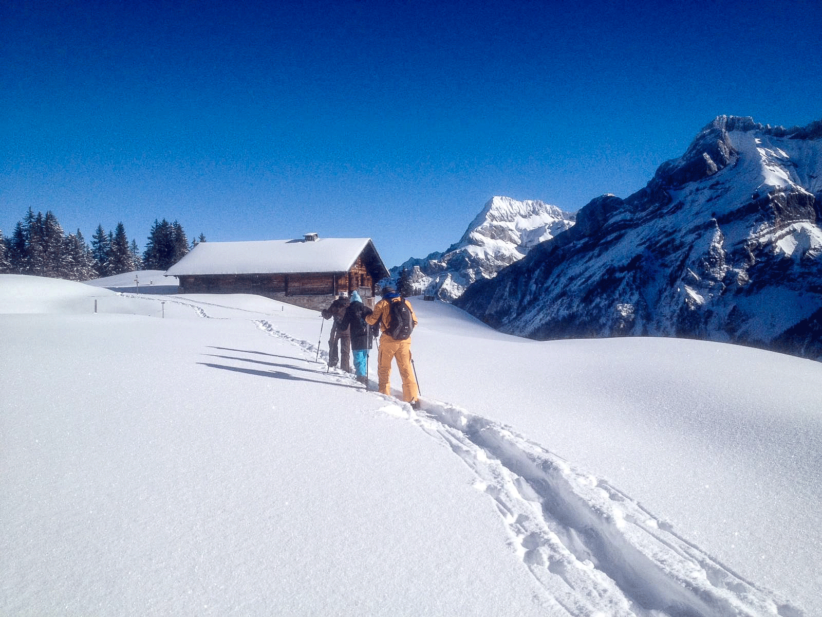 Three people trudging through snow, blue sky, mountain landscape