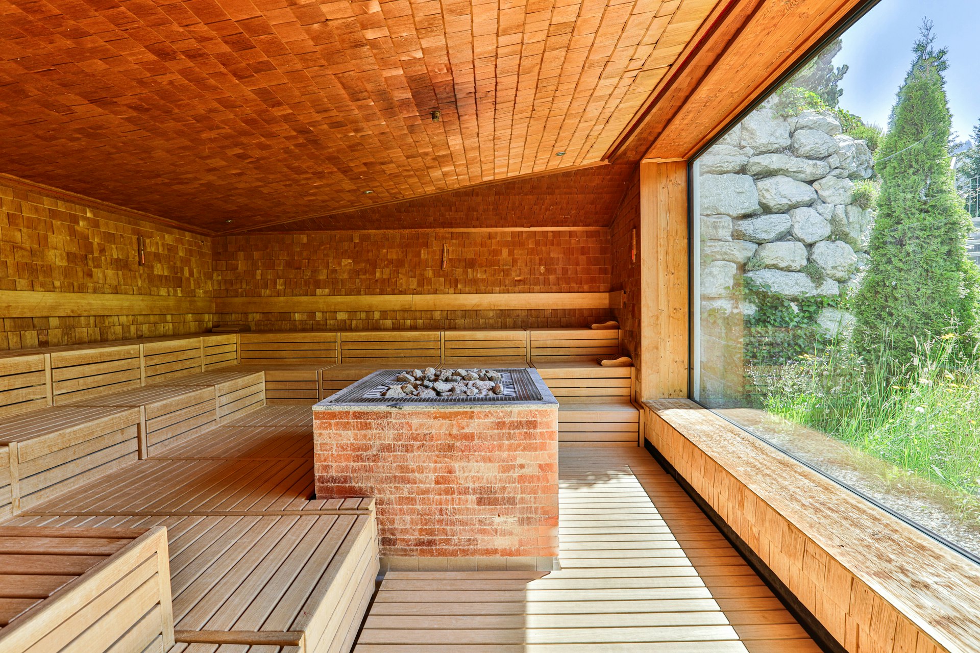 Grosszügige Sauna ganz mit Holz ausgekleidet, Saunaofen in der Mitte, grosses Panoramafenster zum Garten