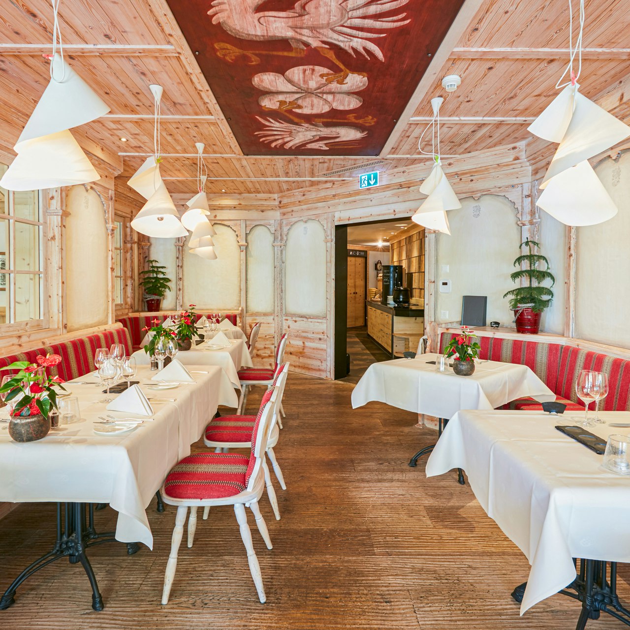 Kranich-Stube im Hotel Ermitage Restaurant mit gedeckten Tischen, oben eine bemalte Holzdecke, Sitzbänke und Sessel mit roten Kissen ausgelegt