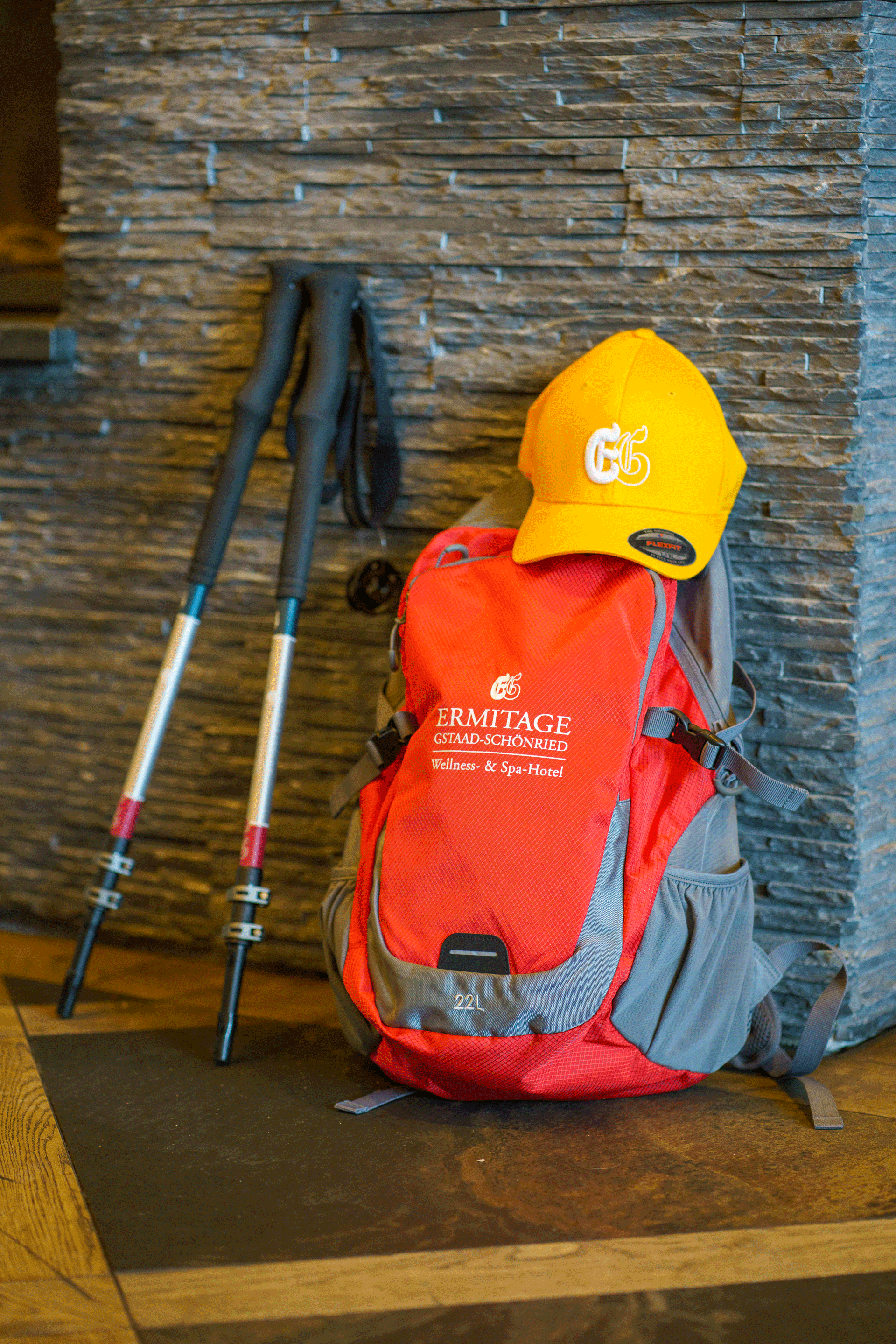 roter Ermitage Rucksack, gelber Ermitage Cap und Ermitage Wanderstöcke vor Steinwand und auf Fussboden im Hotel
