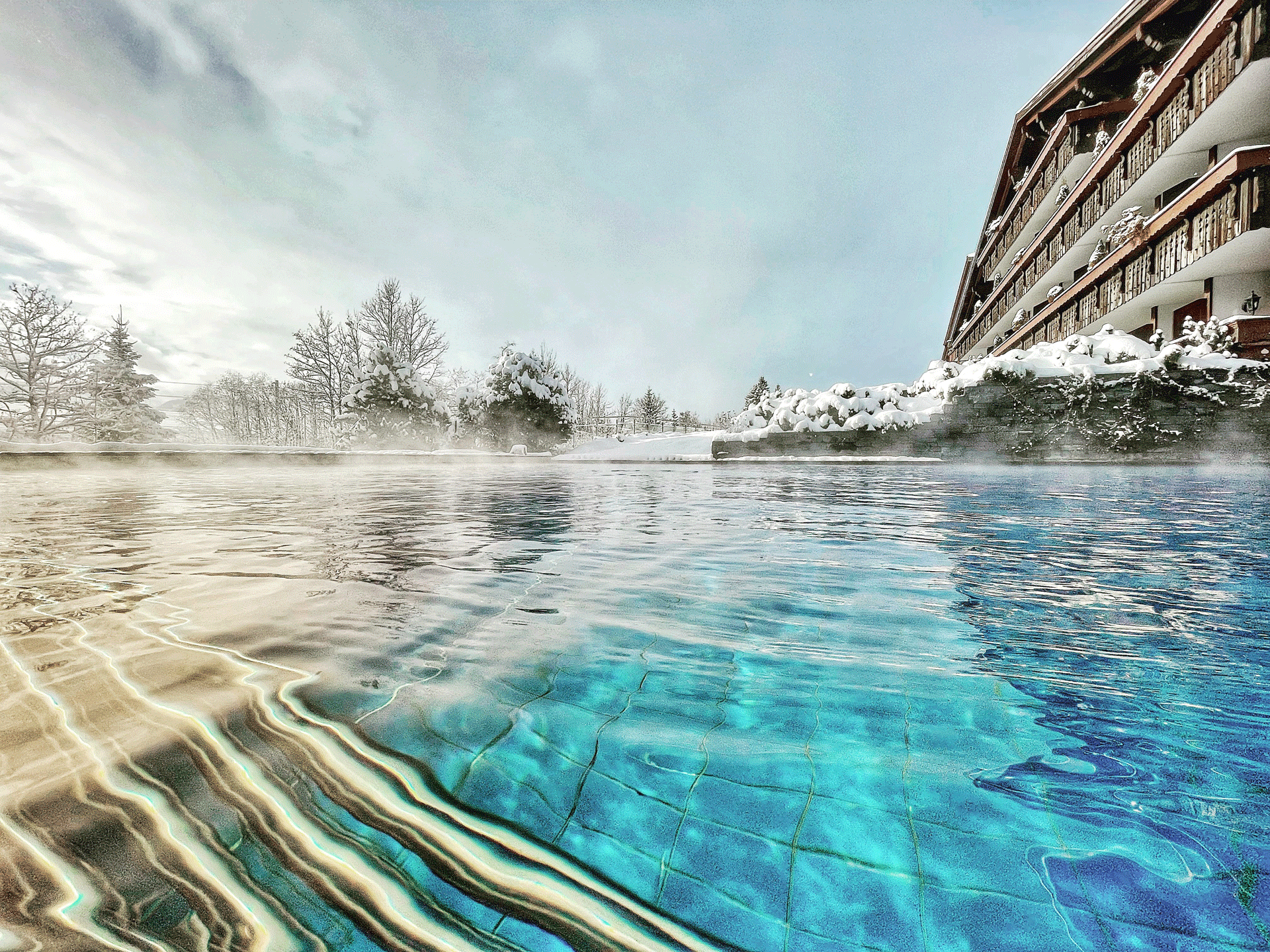Blau schimmerndes Schwimmbecken im Winter, dahinter ein rustikales Holzhaus