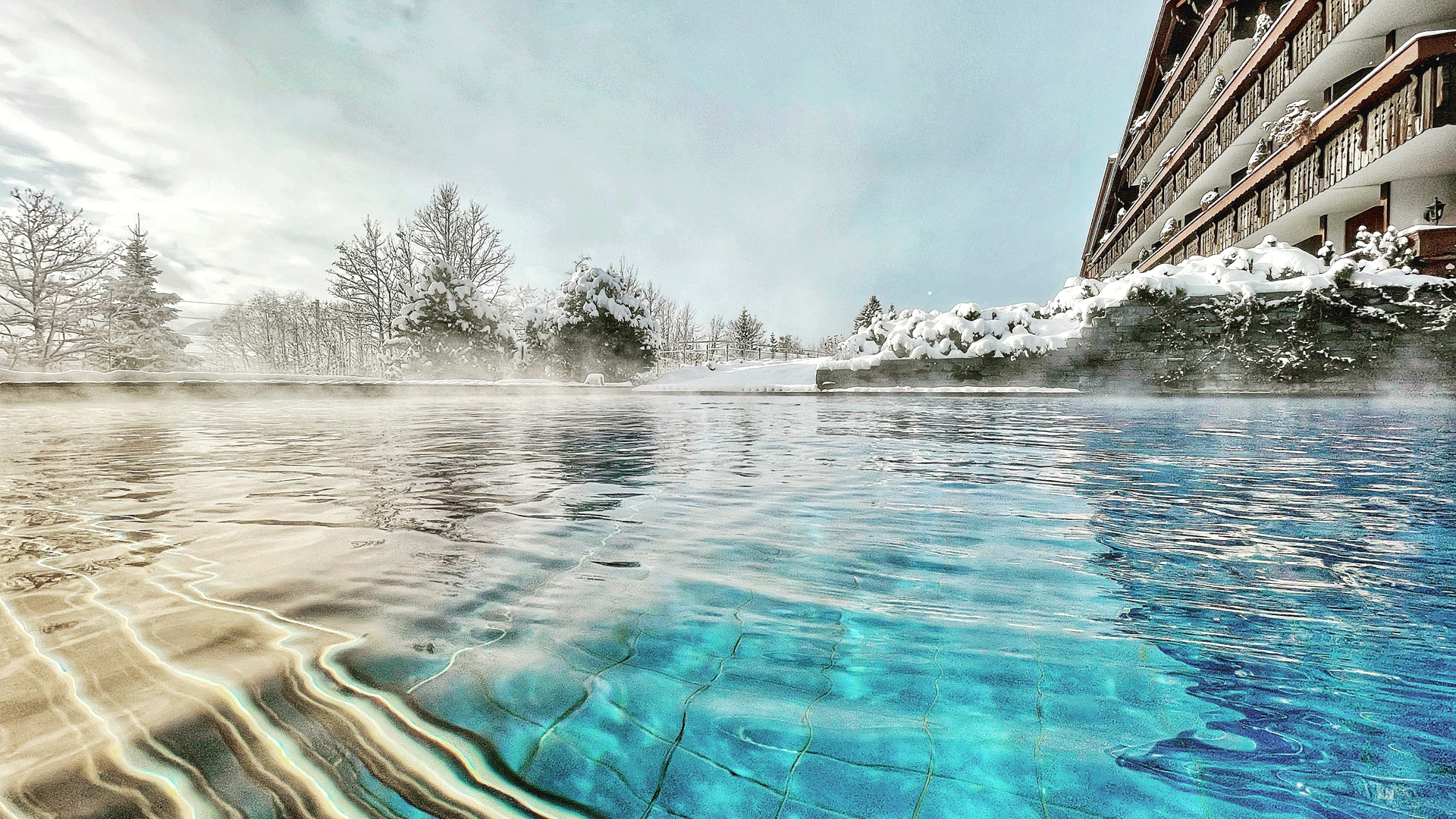 Outdoor pool at the Hotel Ermitage Shimmering blue swimming pool in winter, with a rustic wooden house behind it