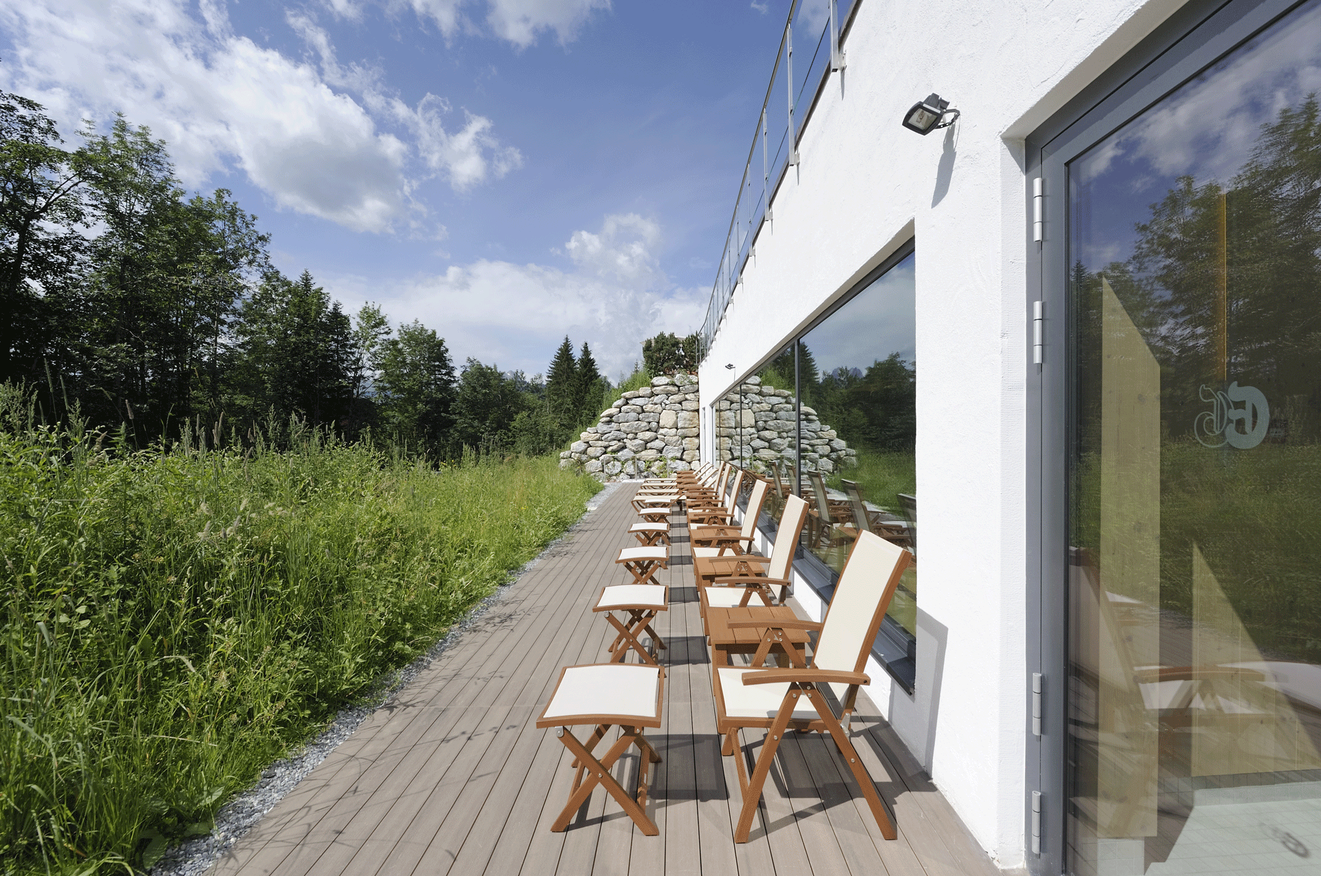 Sonnige Terrasse mit Stühlen an einer Hauswand mit grossen Fenstern, Blick in einen Garten