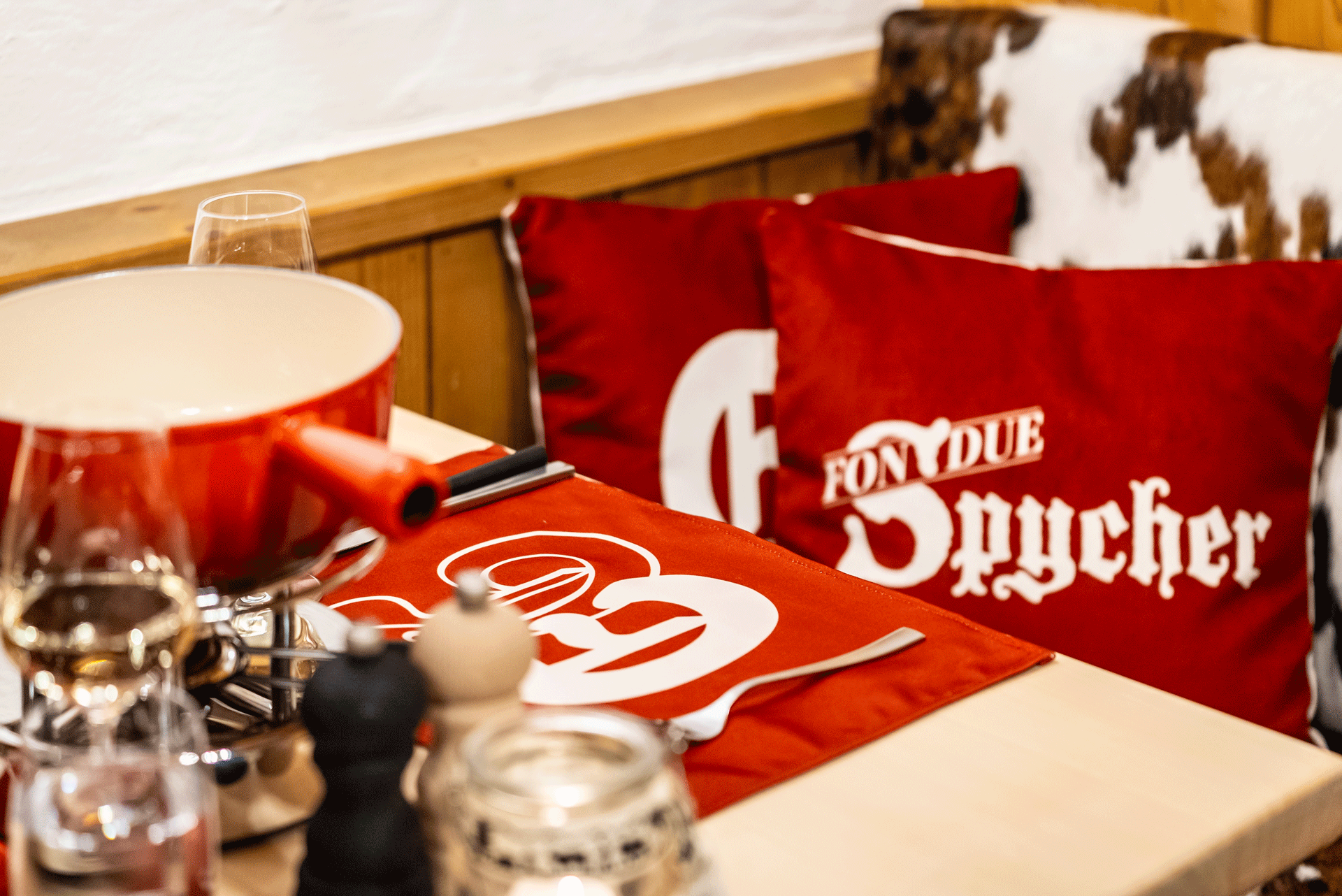 Gedeckter Tisch mit Fondue-Caquelon, rotem Tischset, über dem Stuhl ein Kuhfell, rote Kissen