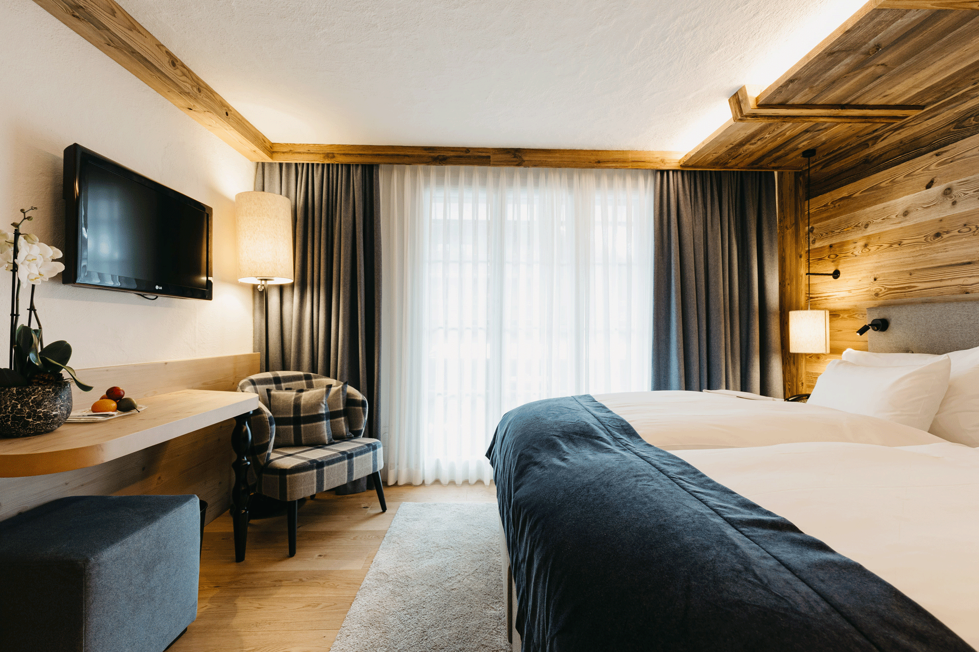 Zimmer mit rustikaler Holzwand, Doppelbett, kleinem Schreibtisch und kariertem Polstersessel
