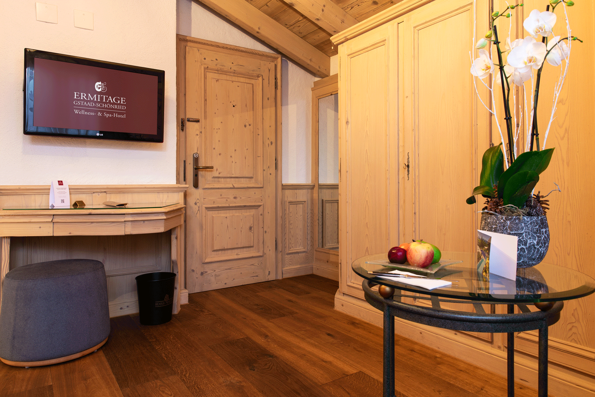 Zimmer mit Dachschräge, Holzboden, Schrank, Glastisch mit Blumen, Arbeitstisch und Flachbildschirm