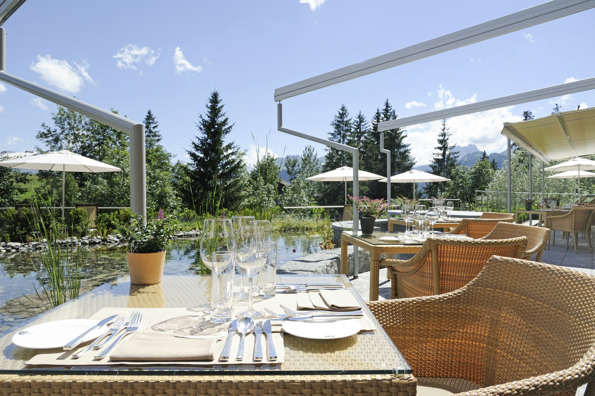 Table dressée avec des meubles en rotin, derrière un étang et des parasols