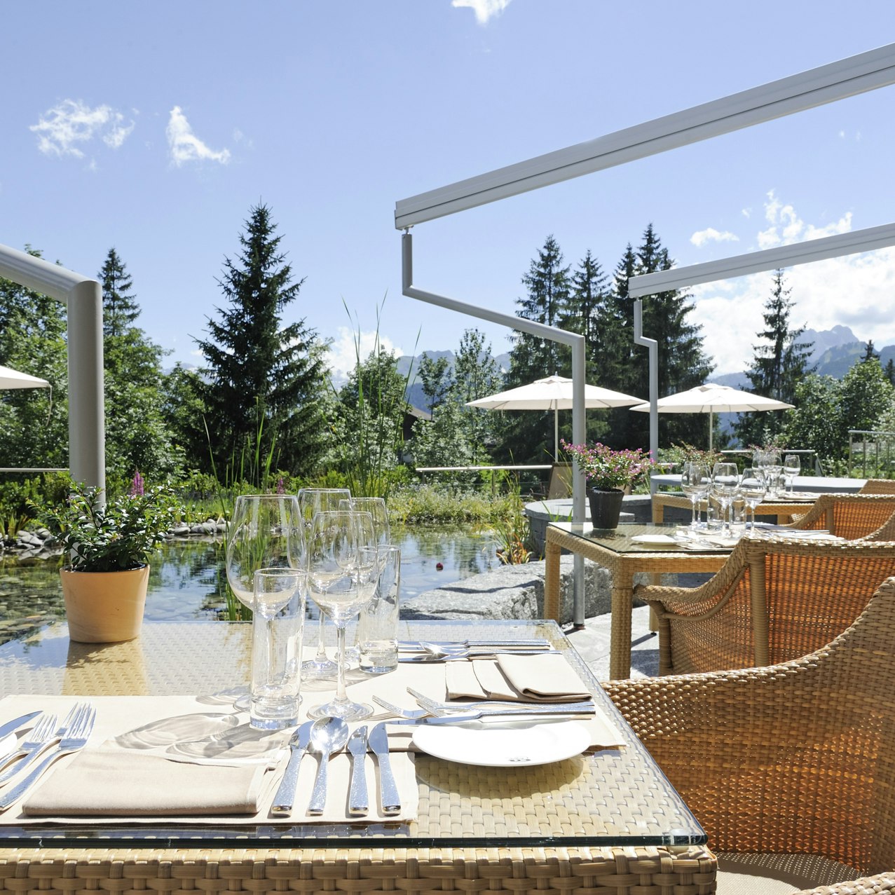 Sonnenterrasse im Hotel Ermitage Gedeckter Tisch mit Rattanmöbeln, dahinter ein Teich und Sonnenschirme