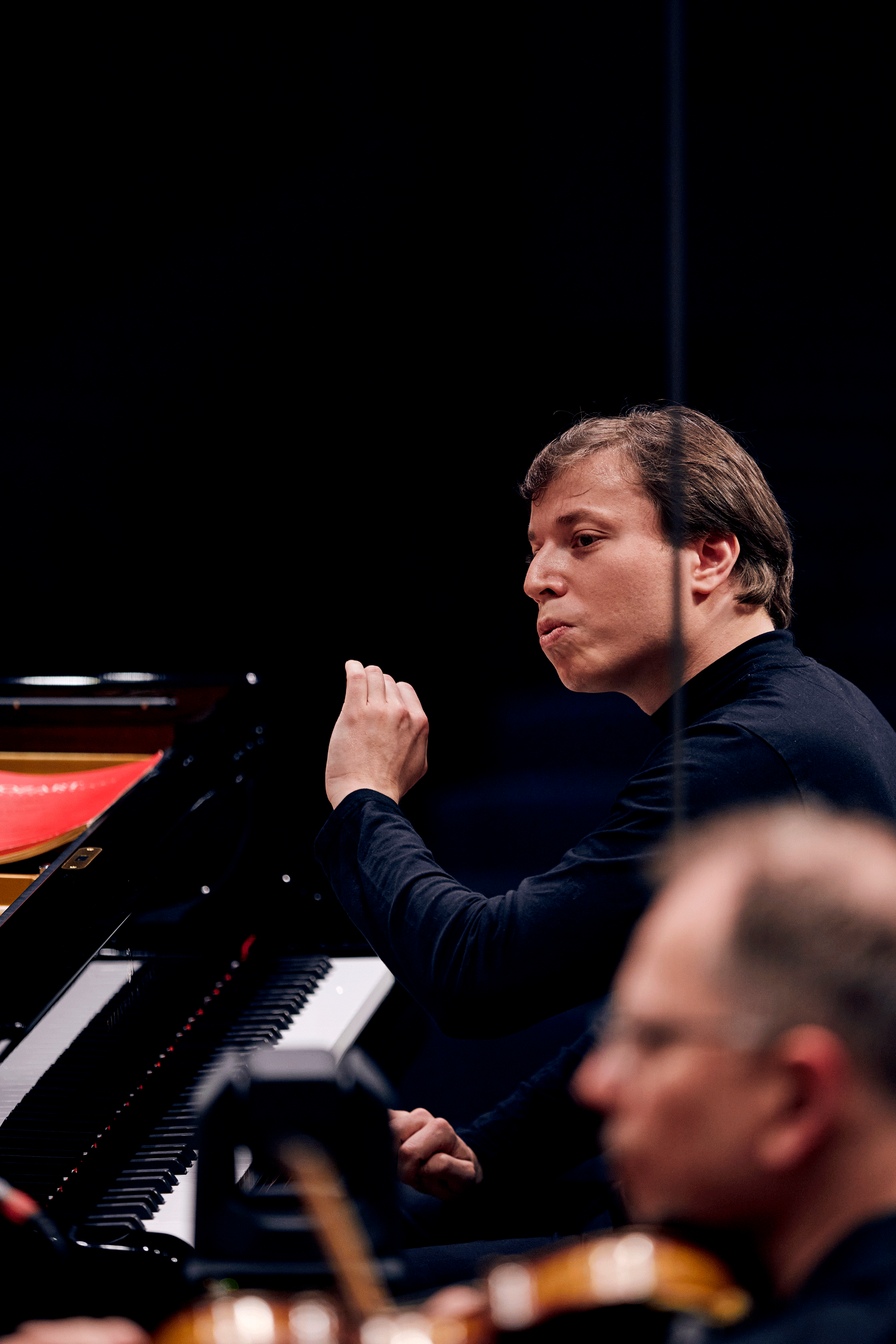 Chef d'orchestre devant un piano, au premier plan dans le flou un violoniste