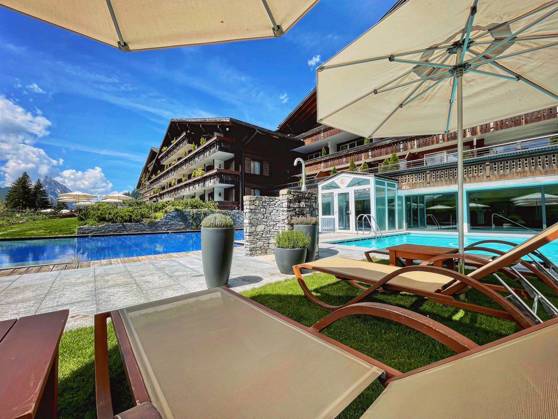 Sun loungers and parasols in front of a dark blue and a light blue pool, behind a large chalet-style house