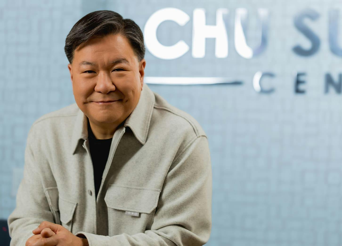 Dr. Chu smiling while sitting in front of his logo