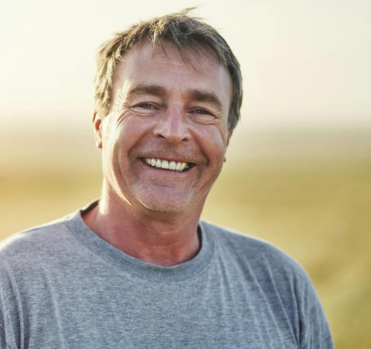 smiling man in grey shirt standing in field with wheat in background