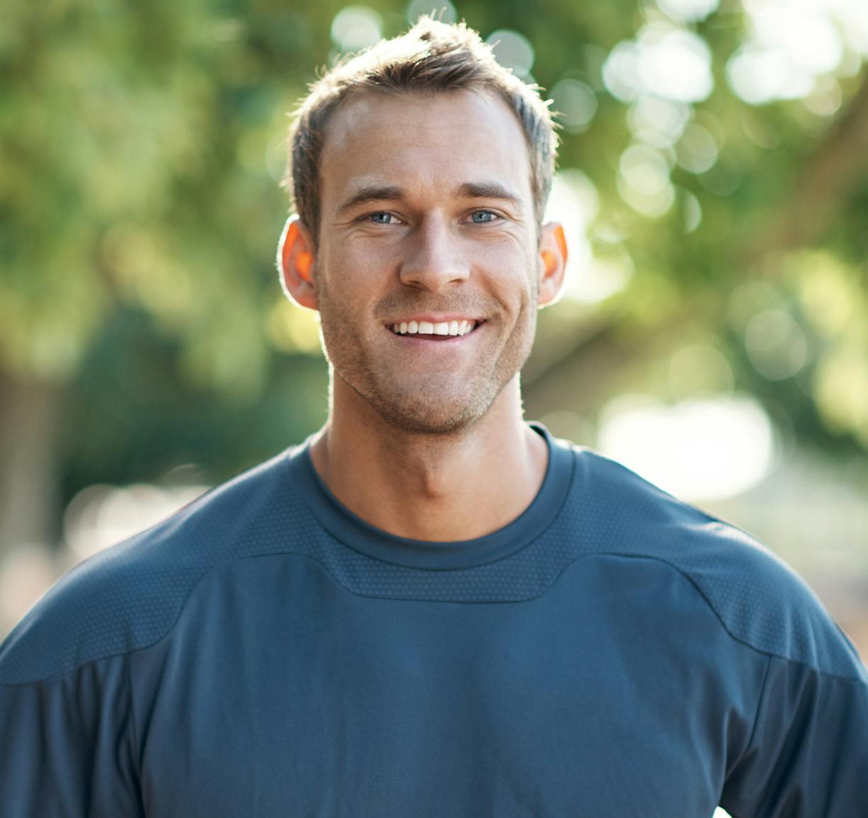 smiling man in blue shirt standing in front of trees