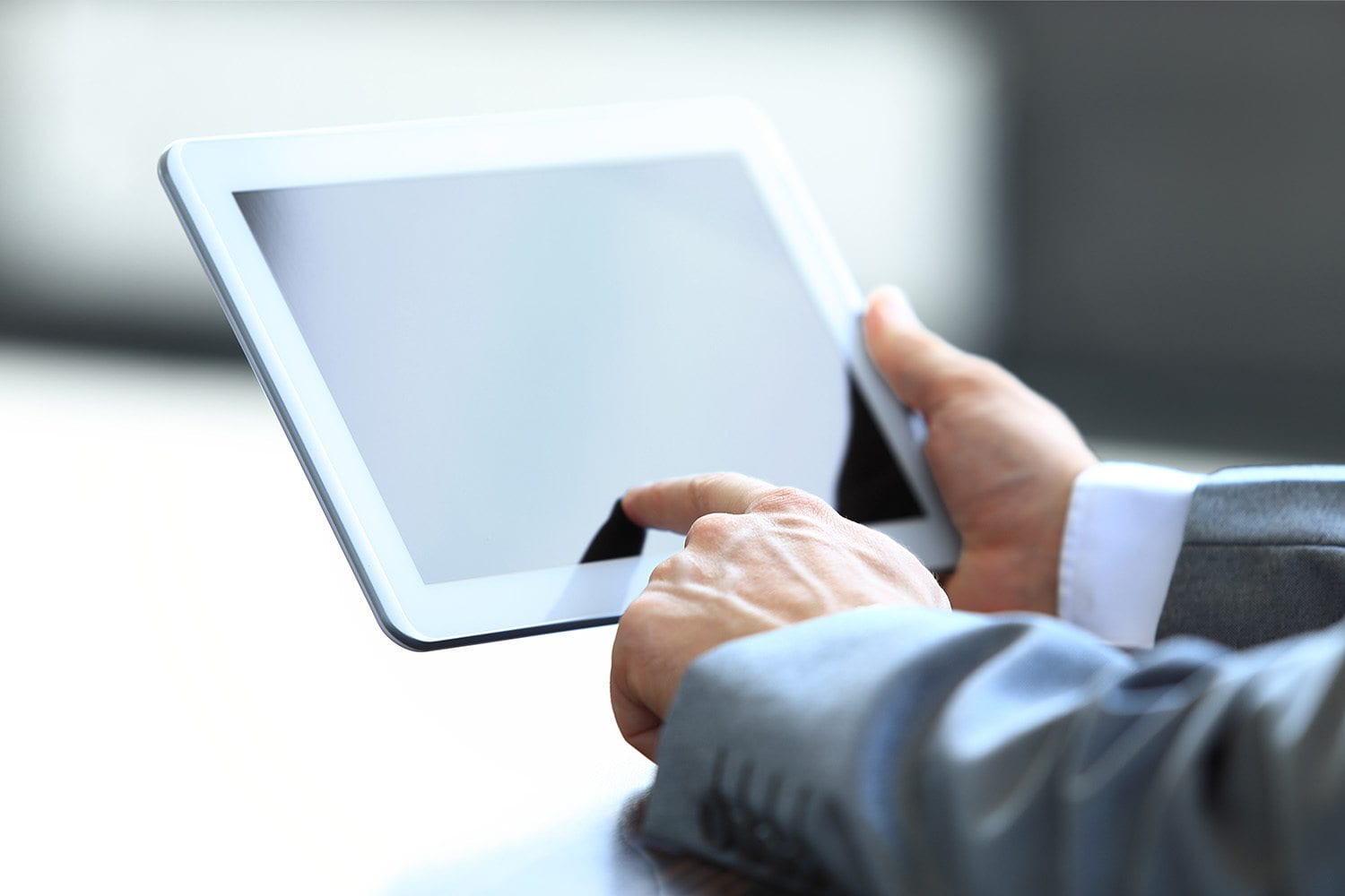 A person in a suit holds a digital tablet with a blank screen, interacting with it using their index finger, set against a plain background.