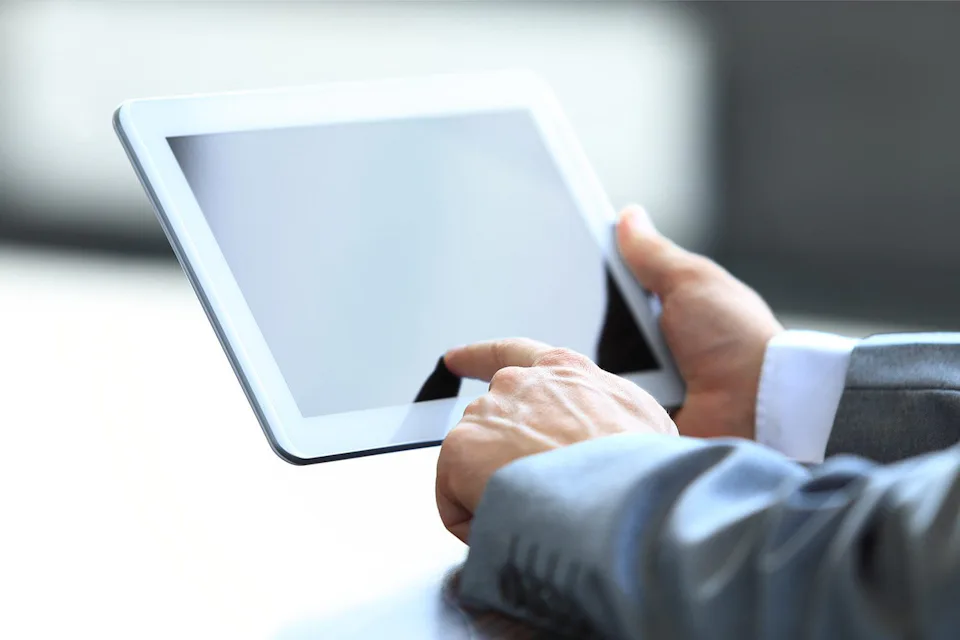 A person in a suit holds a digital tablet with a blank screen, interacting with it using their index finger, set against a plain background.