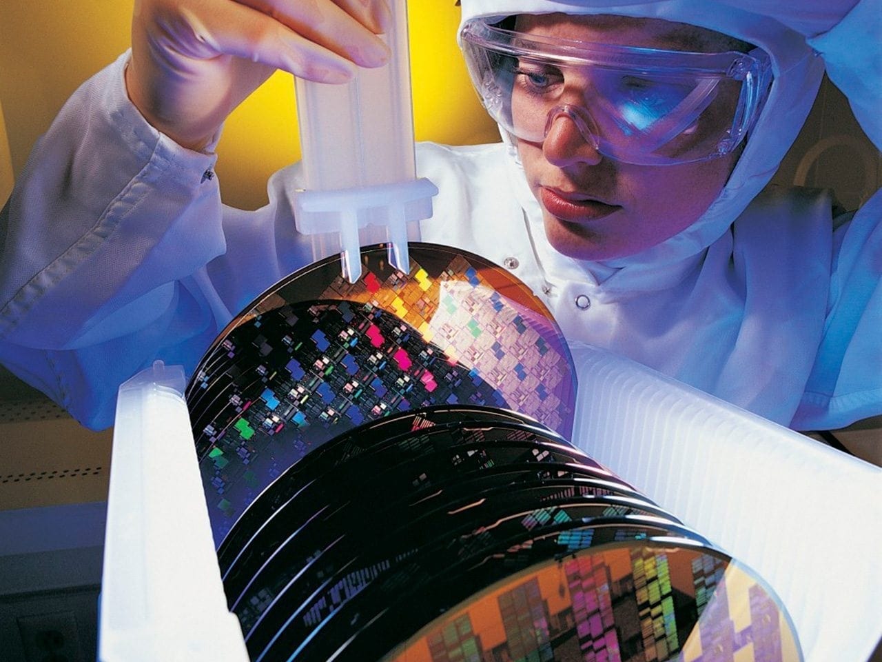 A scientist in protective gear carefully handles a rack of silicon wafers, showcasing intricate microchips, in a laboratory setting, highlighting advanced semiconductor technology.