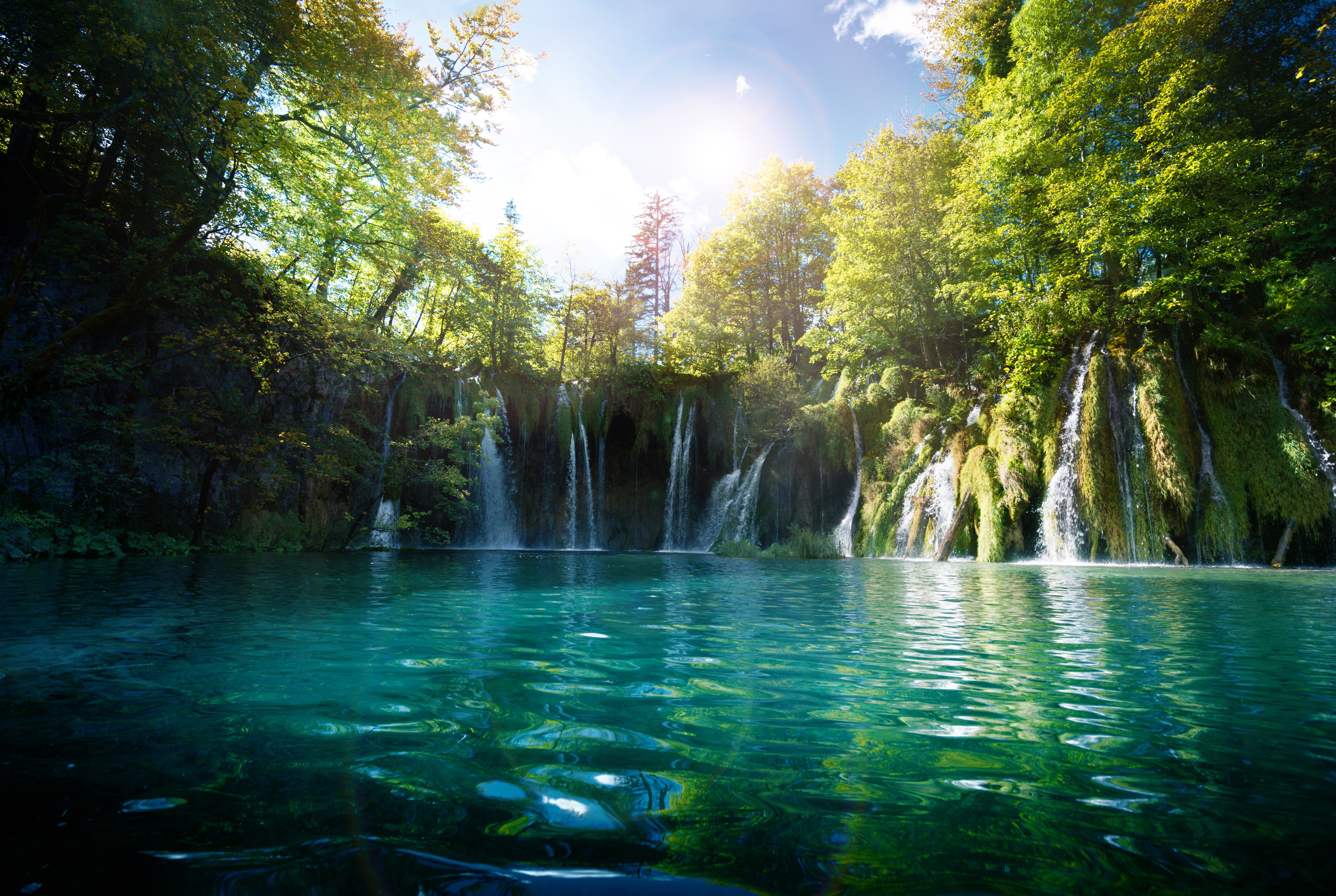 A tranquil waterfall cascades into a clear turquoise lake, surrounded by lush green trees, under a bright, sunny sky.